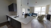 This single-floor space showcases a thoughtfully designed open-concept layout that seamlessly connects the kitchen and living areas. The kitchen features dark cabinetry paired with a light quartz countertop, a stainless steel sink, and a peninsula that extends outward to create a functional breakfast bar. Pendant lighting hangs above the peninsula, adding a modern, stylish touch to the space. The hardwood flooring flows throughout, providing a warm and cohesive look. The living area is generously proportioned and benefits from large windows that invite an abundance of natural light, creating a bright and welcoming ambiance. The high ceilings further enhance the sense of openness and airiness throughout the space. A glimpse into an adjacent bedroom reveals a well-lit, comfortable room with its own access to natural light. The overall design balances practicality with modern aesthetics, making it a versatile and inviting living environment. The neutral color palette across walls and surfaces complements the darker tones of the cabinetry and flooring, resulting in a harmonious and contemporary interior.