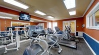 This fitness center is a thoughtfully designed workout facility that offers everything needed for an effective exercise routine. The space is defined by its warm, bold orange accent walls and bright white ceiling with recessed skylights that flood the room with natural light, creating an energizing atmosphere for exercise. Large wall-to-wall mirrors line one side of the room, visually expanding the space and allowing users to monitor their form during workouts. The facility is outfitted with a solid selection of cardio equipment, including treadmills, elliptical trainers, and a stair climber, all arranged to allow comfortable movement between machines. A flat-screen television is mounted on the wall, providing entertainment during workouts. The carpeted flooring adds a degree of comfort underfoot, while the overall layout ensures an open, uncluttered feel. Natural light also enters through windowed doors, contributing to the bright and welcoming environment. The fitness center is a single-floor space designed with both functionality and comfort in mind, making it a practical and inviting destination for daily exercise.
