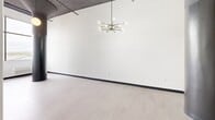 This thoughtfully designed loft-style residence at 480 Myrtle in New Britain, Connecticut offers a compelling blend of industrial character and modern finishes across a single floor. The main living area immediately impresses with its soaring ceilings, exposed structural elements, and clean white walls that create a bright, airy atmosphere. Light-toned flooring throughout the living spaces complements the minimalist aesthetic, while a striking sputnik-style chandelier serves as a bold focal point. A dark-framed entry door adds a dramatic contrast against the crisp white walls, reinforcing the space's industrial-modern identity. Large windows fitted with vertical blinds invite natural light and offer views of the surrounding neighborhood, while an in-unit PTAC system ensures year-round comfort. Two full bathrooms are designed with accessibility in mind, each featuring a tub-shower combination equipped with sturdy grab bars, a shower curtain, and clean white tile. One bathroom is further appointed with a floating vanity, round mirror, and sleek black fixtures, adding a refined, contemporary touch. A hallway connects the various areas of the unit, contributing to a logical and efficient flow. The open, flexible layout of the main room provides ample opportunity for personalized arrangement, making this a versatile and distinctive living space.