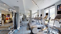This fitness center offers a comprehensive workout environment designed to accommodate a wide range of exercise routines. The space is laid out with a thoughtful arrangement of equipment, featuring a dedicated row of cardio machines—including treadmills, ellipticals, and upright bikes—each equipped with individual screens for entertainment during workouts. Large windows along one wall flood the interior with natural light and provide views of the surrounding greenery, creating an inviting and energizing atmosphere.

On the opposite side of the space, a strength-training zone houses a selection of cable machines and resistance equipment, supplemented by full-length mirrors that visually expand the room and allow users to monitor their form. Structural columns are integrated seamlessly into the layout without disrupting the flow of the space.

Track lighting mounted on the ceiling provides consistent, bright illumination throughout. The floors are covered in a bold, patterned carpet in shades of teal and blue, adding a modern and dynamic aesthetic to the environment. The overall design balances functionality with a polished, contemporary look suited to a professional fitness facility.