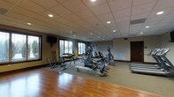 This fitness center offers a comprehensive workout environment designed to accommodate a variety of exercise routines. The space features warm hardwood flooring that transitions to rubberized matting beneath the equipment, providing both durability and safety during workouts. The drop ceiling is fitted with recessed lighting, ensuring consistent, even illumination throughout the room. Large windows along one wall invite natural light and provide views of the surrounding landscape, creating an open and energizing atmosphere. The cardio section is well-stocked with elliptical trainers, a rowing machine, and a stationary bike, while a dedicated free weights area includes a dumbbell rack and weight bench for strength training. A wall-mounted flat-screen television is positioned for easy viewing during workouts. A whiteboard is mounted on one wall, suitable for posting workout schedules or instructions. The neutral-toned walls and dark wood baseboards contribute to a clean, professional aesthetic. A separate door provides additional access or storage options. Overall, the space is thoughtfully organized to support both cardiovascular and strength-based fitness activities.