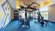 Located at 155 Ames St in Marlborough, MA, this fitness center is a thoughtfully designed, single-room facility that delivers a comprehensive workout environment. The space is defined by its striking architectural details, including a warm wood-paneled ceiling with recessed and ambient lighting that creates an energizing yet welcoming atmosphere. Blue rubber flooring covers the entire area, providing both safety and durability underfoot.

Large floor-to-ceiling windows line multiple walls, flooding the space with natural light and offering views of the surrounding greenery — making workouts feel open and refreshing. A bold geometric mural in blue, yellow, and white adds a vibrant, motivating accent along one wall, complemented by full-length mirrors that enhance the sense of space.

The fitness center is outfitted with professional-grade Hoist equipment, including a multi-functional cable and pulley system at the center of the room. Additional equipment includes a Smith machine with an adjustable bench, a dedicated dumbbell rack with a full range of free weights, kettlebells, resistance bands, and a leg press/cardio machine. A wall-mounted display screen and ceiling fans complete the practical amenities. The layout is well-organized, allowing users to move freely between stations, making this a highly functional space for a full-body training regimen.