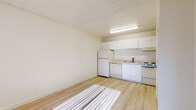 This single-floor unit offers a clean, functional layout with a consistent design language carried throughout its open spaces. Light hardwood-style flooring runs across all areas, reflecting natural sunlight that streams in through vertical-blind-covered windows, creating a warm and airy atmosphere.

The living area is generously proportioned, featuring crisp white walls, a textured ceiling, and a wall-mounted air conditioning unit that provides independent climate control. The entry door opens directly into this main space, which is open and versatile in its use.

The kitchen is efficiently designed and fully equipped with white cabinetry above and below the countertops, a coil-top electric range with oven, a dishwasher, a full-size refrigerator, and a single-basin sink. An overhead range hood and under-cabinet fluorescent lighting provide both function and illumination. Light-toned laminate countertops complement the white cabinetry throughout. An electrical panel is conveniently housed near the kitchen, accessible through a dedicated panel door.

A secondary room, also finished with the same light flooring and white walls, features its own window with vertical blinds and a wall-mounted air conditioning unit, making it suitable as a bedroom or additional living space. Overall, the unit presents a clean, well-maintained environment with practical amenities throughout.