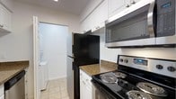 This single-floor space includes a functional kitchen designed with a clean, efficient layout. The kitchen is outfitted with white shaker-style cabinetry extending to the ceiling, providing generous storage throughout. Granite countertops in warm earth tones add a touch of elegance to the workspace. Appliances include a black refrigerator, a stainless steel over-the-range microwave, and a black electric range, offering a practical and modern cooking environment. Light-toned ceramic tile flooring runs through the kitchen, contributing to a bright and clean aesthetic. A glimpse into the adjacent hallway reveals a stacked washer and dryer unit, indicating convenient in-unit laundry facilities. The white walls and recessed lighting keep the space feeling open and well-illuminated. The overall design balances functionality with a neutral, contemporary style, making the kitchen a hardworking central feature of this single-story space.