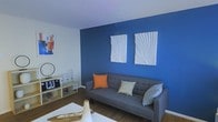 This single-floor space showcases a striking and contemporary interior design that immediately draws the eye. The living area serves as the centerpiece, featuring a bold cobalt blue accent wall that creates a vivid focal point against the surrounding neutral white walls and ceiling. Two white textured sculptural art panels are mounted on the blue wall, adding depth and visual interest to the space. A sleek gray sofa with a tufted design anchors the seating area, accented by colorful throw pillows in orange and sage tones that complement the room's vibrant color palette. Warm hardwood flooring runs throughout, adding a natural contrast to the cool blue tones of the walls. A low-profile media console along the side wall provides functional storage and display space, decorated with minimalist accessories and small décor items. A white coffee table in the foreground ties the room together, while a piece of colorful abstract artwork on the adjacent wall adds an artistic touch. The overall ambiance of the space is modern, lively, and carefully styled, balancing bold color choices with clean lines and purposeful furnishings to create a cohesive and welcoming environment.