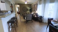 This single-floor residence offers a thoughtfully designed open-concept layout that flows naturally from the dining area into a comfortable living room. Rich hardwood-style flooring runs throughout, lending warmth and continuity to the space. Crown molding adds a refined architectural touch along the ceilings, while a ceiling fan with a decorative light fixture serves as a functional centerpiece overhead.

The living area is anchored by a cozy seating arrangement, featuring a sofa and accent chairs in dark wood frames with neutral upholstery. A dark wood side table with a classic table lamp and carefully placed greenery enhance the inviting atmosphere. Framed artwork adorns the walls, complementing the earthy, olive-toned accent wall that adds depth and character to the room.

Large windows dressed in white blinds and sheer curtains allow natural light to filter in, brightening the interior throughout the day. The dining area, positioned adjacent to the living space, includes bar-height seating along one side, ideal for casual dining or entertaining.

A hallway visible in the background connects to additional rooms, including what appears to be a bedroom, hinting at a practical and well-organized floor plan. Overall, the space exudes a warm, contemporary ambiance with tasteful décor and a cohesive design aesthetic.