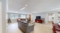 Located at 341 S Ellington Pkwy in Lewisburg, Tennessee, this single-story community clubhouse offers a well-organized and functional layout designed for social gatherings and relaxation. The interior is finished with light wood-look flooring throughout, neutral gray walls, and flush-mounted fluorescent lighting that keeps the space bright and welcoming.

The expansive living room serves as the heart of the clubhouse, comfortably accommodating multiple seating arrangements with leather sofas and a glass-top coffee table. A flat-screen television on a wooden media console anchors one end of the room, while a dining area with a full table and chairs sits near a bank of windows that allow natural light to fill the space.

The kitchen is outfitted with white cabinetry, light granite-style countertops, and white appliances including a range, microwave, and full-size refrigerator — all set on the same wood-look flooring for a cohesive feel. A dedicated entrance provides easy access to the building.

Outside, a large rectangular swimming pool is the centerpiece of the outdoor area, surrounded by a concrete deck lined with lounge chairs for sunbathing. A chain-link fence encloses the pool area, and mature trees provide a natural green backdrop. A small storage shed and additional seating complete the outdoor amenity space.