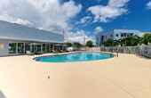 Located at 13081 SW 242nd St in Princeton, Florida, this outdoor amenity space is centered around a generously sized oval swimming pool with crystal-clear blue water. The pool is finished with a smooth, light-toned deck that provides ample space for lounging and recreation. A secure wrought iron fence encloses the pool perimeter, ensuring safety while maintaining an open, airy feel with views of surrounding palm trees and lush tropical landscaping.

Adjacent to the pool, a well-appointed sun deck features multiple padded chaise lounges paired with large shade umbrellas, offering comfortable options for relaxation in the South Florida sun. The deck's neutral-toned surface extends generously around the pool, allowing for easy movement and gathering.

A modern single-story clubhouse building with a distinctive blue-gray stucco exterior borders the pool area. Its floor-to-ceiling glass facade and covered breezeway seamlessly connect the interior to the outdoor space, flooding both areas with natural light. The building's clean, contemporary architectural lines complement the resort-style atmosphere of the outdoor amenity area.

The overall ambiance is bright and inviting, enhanced by the tropical Florida climate, swaying palms, and open skies. This well-maintained outdoor space is ideally suited for community leisure and social activities.