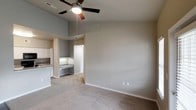 This single-floor residence offers a clean, open layout designed for comfortable living. The main living area immediately draws the eye upward with its vaulted ceilings, which lend a sense of openness and airiness to the space. A ceiling fan with a built-in light fixture is centrally mounted, providing both air circulation and ambient lighting. The living area is finished with wall-to-wall plush carpeting in a neutral tone, complementing the soft gray walls and crisp white baseboards throughout.

Flowing seamlessly from the living area, the kitchen is partially open to the main space, creating a connected feel. The kitchen features granite countertops with attractive natural veining, white cabinetry, and a full-size refrigerator, offering a functional and visually cohesive cooking environment. A dedicated built-in desk or utility counter area is also visible adjacent to the kitchen, providing additional workspace or storage potential.

A doorway leads further into the home, suggesting additional rooms beyond. The overall color palette — soft grays, warm whites, and neutral tones — creates a calm and inviting ambiance throughout. The space presents a practical yet comfortable layout suited for everyday living.