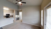 This single-floor residence offers a clean, open layout designed for comfortable living. The main living area immediately draws the eye upward with its vaulted ceilings, which lend a sense of openness and airiness to the space. A ceiling fan with a built-in light fixture is centrally mounted, providing both air circulation and ambient lighting. The living area is finished with wall-to-wall plush carpeting in a neutral tone, complementing the soft gray walls and crisp white baseboards throughout.

Flowing seamlessly from the living area, the kitchen is partially open to the main space, creating a connected feel. The kitchen features granite countertops with attractive natural veining, white cabinetry, and a full-size refrigerator, offering a functional and visually cohesive cooking environment. A dedicated built-in desk or utility counter area is also visible adjacent to the kitchen, providing additional workspace or storage potential.

A doorway leads further into the home, suggesting additional rooms beyond. The overall color palette — soft grays, warm whites, and neutral tones — creates a calm and inviting ambiance throughout. The space presents a practical yet comfortable layout suited for everyday living.