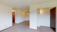 This single-floor residence offers a straightforward, functional layout designed for comfortable everyday living. The main living area is finished with wall-to-wall neutral carpet and crisp white walls accented by dark baseboards, creating a clean and simple aesthetic throughout. A ceiling fan with a light fixture is centrally mounted overhead, providing both air circulation and ambient lighting to the space.

The open floor plan allows for flexible furniture arrangement, with the living area flowing naturally toward adjoining rooms. Multiple doorways branch off from the main space — one leading to what appears to be a hallway or additional room with warm-toned wood-style finishes, and another leading to a secondary area with its own wall-mounted light source. A partial wall divider adds a subtle architectural element that helps define distinct zones within the open layout without fully closing off the space.

Overall, the residence presents a neutral, move-in-ready canvas with an uncluttered design that allows for easy personalization. The straightforward layout, consistent flooring, and practical ceiling fan make this a functional and adaptable single-floor living space suited to a variety of lifestyles.