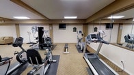 This fitness center is a compact yet comprehensively outfitted space designed to support a full range of exercise routines. The room features a neutral color palette with warm beige walls and carpeted flooring, creating a clean and focused workout atmosphere. Recessed flat panel lighting illuminates the space evenly from above, ensuring excellent visibility throughout.

The cardio equipment selection includes a treadmill, an elliptical trainer, a recumbent stationary bike, and a rowing-style machine, each positioned on protective rubber mats. For strength training, a multi-station cable and weight machine is available alongside a flat workout bench, catering to a variety of resistance exercises. A flat-screen television is mounted on the wall, providing entertainment during workouts.

A large wall mirror spans a significant portion of the room, effectively amplifying the sense of space and allowing users to monitor their form during exercise. A central structural column is present in the room, which slightly divides the floor plan while maintaining an open and accessible feel. Informational signage is posted on the walls, offering guidance on equipment use and facility rules. Overall, the fitness center is a practical and well-maintained space suited for a thorough daily workout.