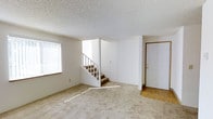 This two-story residence offers a straightforward and functional layout designed for comfortable everyday living. The main level centers around a spacious living area finished with wall-to-wall carpet in a warm beige tone, providing a soft and practical underfoot surface. White walls throughout create a clean, neutral backdrop that lends itself well to a variety of furnishing styles.

A centrally positioned interior staircase with a white-painted wood railing connects the main level to the upper floor, serving as a natural dividing point within the open living space. Near the entry, the flooring transitions from carpet to hard surface, indicating a defined entryway area just inside the front door, which features warm wood-toned trim detailing.

Natural light enters the main living area through a large window fitted with vertical blinds, casting bright light across the carpeted floor. The textured popcorn ceiling is consistent throughout, adding a classic residential character to the space. The overall ambiance is clean, bright, and understated, offering a neutral foundation that is well-suited for personalization and comfortable daily use across both levels.