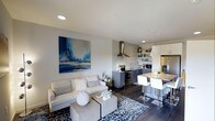 This thoughtfully designed single-floor space showcases a seamless open-concept layout that effortlessly connects the living and kitchen areas. The living room is anchored by a plush light-toned sectional sofa, complemented by a patterned area rug and a striking large-format blue abstract artwork that adds a bold visual focal point to the neutral gray walls. Recessed lighting illuminates the space, enhancing its clean, modern aesthetic.

The kitchen is a standout feature, boasting dark gray cabinetry paired with a light subway tile backsplash and open shelving that provides both function and style. A stainless steel range hood crowns the cooking area, which is equipped with stainless steel appliances including a refrigerator and electric range. Pendant lights hang above a granite-topped breakfast bar with white bar seating, creating a casual dining area that bridges the kitchen and living space.

Dark hardwood flooring flows throughout, providing a warm contrast to the lighter furnishings and cabinetry. The overall ambiance is contemporary and cohesive, with a carefully curated color palette of grays, whites, and blues that gives the space a polished, inviting character suited for both everyday living and entertaining.