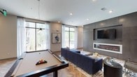 Located at 3310 Women's Club Dr in Raleigh, NC, this single-story amenity lounge offers a sophisticated and contemporary gathering space designed for both relaxation and entertainment. The open layout is anchored by a full-size pool table positioned beneath a sleek pendant light, making it an immediate focal point upon entering. Adjacent to the billiards area, a plush navy sectional sofa is arranged around a patterned area rug, creating an inviting lounge zone centered on a wall-mounted flat-screen television. A modern linear fireplace, set within a floor-to-ceiling concrete-paneled accent wall, adds warmth and architectural depth to the space. The herringbone-patterned hardwood flooring lends a refined, upscale character throughout. Large floor-to-ceiling windows dressed with sheer white curtains flood the interior with abundant natural light while framing views of the surrounding greenery. The neutral color palette of warm grays, whites, and deep blues contributes to a polished yet comfortable ambiance. Recessed lighting throughout the ceiling complements the natural light, ensuring the space remains bright and well-illuminated at all times.