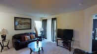 This single-floor space centers around a well-designed living room that balances comfort and functionality. The living room is finished with wall-to-wall carpeting and neutral-toned walls, creating a warm and inviting atmosphere. A sliding glass door, dressed with dark curtain panels, allows natural light to filter into the room while providing access to an outdoor area. A side window with blinds further supplements the natural lighting. The space accommodates a dark leather sectional sofa accented with decorative pillows in warm tones, paired with a round glass-top coffee table that adds a touch of elegance. A large framed artwork adorns one wall, contributing a refined aesthetic to the room. On the opposite side, a flat-screen television is mounted on a wooden media stand, serving as a practical entertainment focal point. A floor lamp positioned near the seating area provides additional ambient lighting. The overall layout is efficient and thoughtfully arranged, making good use of the available space while maintaining an open, uncluttered feel throughout the living area.