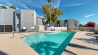 Located at 8560 W Peoria Ave in Peoria, Arizona, this outdoor pool area is a well-appointed recreational space designed for enjoying the sunny Southwest climate. The centerpiece is a generously sized swimming pool with crystal-clear, turquoise water, framed by a smooth concrete pool deck that provides ample space for lounging. Multiple padded chaise lounge chairs and outdoor seating arrangements are positioned around the pool, offering comfortable spots to relax and take in the surroundings.

The area is enclosed by a combination of wrought iron fencing and perimeter walls, providing both security and privacy. Mature eucalyptus and palm trees rise above the enclosure, casting natural shade and adding a lush, desert-oasis feel to the space. Vibrant seasonal plantings, including flowering bougainvillea, add pops of color against the neutral stucco exterior of the adjacent multi-story buildings.

A covered gazebo or ramada structure is visible along one side of the pool deck, offering shaded seating and gathering space. The surrounding buildings feature a contemporary stucco exterior in neutral gray and blue tones with multi-pane windows. The entire space is bathed in abundant natural light beneath the characteristic bright blue Arizona sky, creating an inviting and refreshing outdoor retreat.