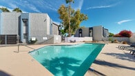 Located at 8560 W Peoria Ave in Peoria, Arizona, this outdoor pool area is a well-appointed recreational space designed for enjoying the sunny Southwest climate. The centerpiece is a generously sized swimming pool with crystal-clear, turquoise water, framed by a smooth concrete pool deck that provides ample space for lounging. Multiple padded chaise lounge chairs and outdoor seating arrangements are positioned around the pool, offering comfortable spots to relax and take in the surroundings.

The area is enclosed by a combination of wrought iron fencing and perimeter walls, providing both security and privacy. Mature eucalyptus and palm trees rise above the enclosure, casting natural shade and adding a lush, desert-oasis feel to the space. Vibrant seasonal plantings, including flowering bougainvillea, add pops of color against the neutral stucco exterior of the adjacent multi-story buildings.

A covered gazebo or ramada structure is visible along one side of the pool deck, offering shaded seating and gathering space. The surrounding buildings feature a contemporary stucco exterior in neutral gray and blue tones with multi-pane windows. The entire space is bathed in abundant natural light beneath the characteristic bright blue Arizona sky, creating an inviting and refreshing outdoor retreat.
