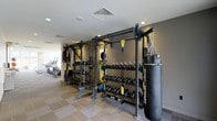 This modern fitness facility is thoughtfully designed to accommodate a wide variety of training styles and fitness goals. The space is anchored by a robust functional training rig system, complete with suspension trainers and pull-up bars, providing a versatile platform for bodyweight and resistance-based workouts. Flanking the rig are neatly organized dumbbell racks stocked with a comprehensive range of weights, alongside kettlebells and barbells with weight plates, ensuring that free weight training is fully catered for. A heavy punching bag adds a dedicated space for combat-style conditioning and cardio training. The flooring features a practical tiled surface with rubberized matting in key workout zones, balancing durability with comfort and safety. Recessed and flush-mounted lighting keeps the space well-lit throughout, while the neutral grey and white color palette lends a clean, professional atmosphere. In the background, a dedicated cardio zone is visible, housing machines such as ellipticals and stationary bikes. The overall layout is open and efficiently organized, making navigation and equipment access straightforward for users of all fitness levels.