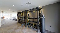 This modern fitness facility is thoughtfully designed to accommodate a wide variety of training styles and fitness goals. The space is anchored by a robust functional training rig system, complete with suspension trainers and pull-up bars, providing a versatile platform for bodyweight and resistance-based workouts. Flanking the rig are neatly organized dumbbell racks stocked with a comprehensive range of weights, alongside kettlebells and barbells with weight plates, ensuring that free weight training is fully catered for. A heavy punching bag adds a dedicated space for combat-style conditioning and cardio training. The flooring features a practical tiled surface with rubberized matting in key workout zones, balancing durability with comfort and safety. Recessed and flush-mounted lighting keeps the space well-lit throughout, while the neutral grey and white color palette lends a clean, professional atmosphere. In the background, a dedicated cardio zone is visible, housing machines such as ellipticals and stationary bikes. The overall layout is open and efficiently organized, making navigation and equipment access straightforward for users of all fitness levels.