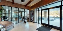 This single-floor lobby showcases a compelling fusion of industrial and contemporary design elements. Exposed red brick walls and dark wood ceiling beams establish a raw, architectural character, while large black-framed glass doors and floor-to-ceiling windows flood the space with natural light and offer clear views of the surrounding exterior.

The entry area features large-format light stone tile flooring, transitioning to warm hardwood in the corridor beyond. A dramatic floor-to-ceiling metal-paneled fireplace serves as a striking focal point in the lounge area, surrounded by plush upholstered seating, including barrel chairs and a decorative side table, creating an inviting gathering space.

Curated abstract artwork is displayed throughout, adding bold color and visual interest against the textured wall panels and brick backdrops. A glass-partitioned room adjacent to the main entry area provides a dedicated workspace or meeting area, visible yet separate from the lobby.

The corridor leading deeper into the building features a patterned area rug over hardwood flooring, adding warmth and direction. The overall ambiance is polished yet approachable, balancing modern sophistication with welcoming, residential comfort.