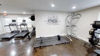 This single-level fitness space is thoughtfully designed for serious home workouts. The room features a durable polished concrete floor that provides a sturdy, easy-to-clean surface well-suited for heavy exercise equipment. Recessed lighting in the ceiling ensures the space is well-lit throughout, while a window along one wall allows natural light to filter in, adding an airy quality to the room.

A bold fitness-themed word art mural serves as the focal point of the main wall, featuring motivational terms centered around the word "FITNESS," adding an energetic and purposeful atmosphere to the space. The neutral, light gray walls complement the industrial feel of the flooring and equipment.

The room is equipped with a treadmill and a rowing machine positioned near the window, offering cardio options with a view. A Rogue-branded lifting platform with a loaded barbell occupies the center of the room, ideal for strength training. A multi-function squat and power rack stands along the opposite wall, rounding out the strength equipment offerings. Overall, the space strikes a balance between functional design and motivating aesthetics.