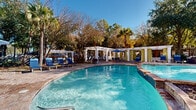 This expansive outdoor amenity area centers around a generously sized freeform swimming pool with clear, blue-tinted water and a smooth stamped concrete and stone surround. The pool's organic, curved shape adds a resort-like character, with easy-access entry steps and handrails at multiple points. Adjacent to the main pool is a connected shallow wading area, ideal for relaxing at the water's edge.

A striking white pergola structure lines one side of the pool deck, providing shaded lounging areas equipped with blue chaise lounges, patio seating, market umbrellas, and private curtained cabanas draped in green — offering both comfort and a touch of elegance. The open-air design of the pergola allows natural light to filter through while providing relief from the sun.

The pool area is flanked by a charming single-story clubhouse pavilion featuring a pitched shingle roof, ceiling fan, large glass-paned doors, and an open porch with additional seating — seamlessly blending indoor and outdoor living. Mature palm trees, ornamental shrubs, cycads, and seasonal flowering plants create lush, layered landscaping throughout the grounds, enhancing the tropical ambiance. The entire space is enclosed by decorative iron fencing, offering both privacy and security under brilliantly clear skies.