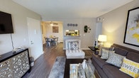 This single-floor residence offers a comfortable and thoughtfully designed living environment within an attractively landscaped community. The exterior setting is a standout feature, with a generously sized outdoor swimming pool framed by a spacious sun deck, lounge seating, and mature trees that provide both shade and a sense of privacy. The surrounding buildings feature warm wood-toned architectural details and multi-level balconies, contributing to a relaxed, resort-like atmosphere under open blue skies.

Inside, the open-plan living and dining area is finished with rich, dark hardwood-style flooring that runs throughout, creating a cohesive and inviting ambiance. The living room is well-proportioned, accommodating comfortable seating arranged around a glass-top coffee table. A pass-through opening connects the space to the kitchen, enhancing the sense of openness and allowing natural interaction between the cooking and living areas. The dining area, visible from the living room, features a ceiling fan and ample space for a full dining set. Neutral wall tones throughout keep the interior bright and versatile, while decorative touches lend the space a curated, homey character.