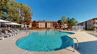 Located at 5600 Fountain Grove Circle in Fayetteville, North Carolina, this outdoor amenity area serves as the centerpiece of the Fountain Grove community. The space is anchored by a generously sized freeform swimming pool with crystal-clear blue water, gentle curvilinear edges, and multiple entry points via stainless steel handrails and steps. The pool area accommodates varying depths, as indicated by clearly marked depth signage, ensuring safety for all users.

Surrounding the pool is a spacious concrete sundeck lined with numerous lounge chairs and chaise lounges, offering ample space for relaxation under the open sky. A covered gazebo on one side of the deck provides shaded seating, while a modern white pergola with a built-in lounge set and fire table offers an additional gathering area near the gated entrance.

The adjacent single-story clubhouse building features a covered porch with ceiling fans and columns, opening directly onto the pool deck. Lounge seating is arranged along the building's exterior, creating a seamless indoor-outdoor connection. A separate utility structure near the entrance provides additional support facilities.

The entire area is enclosed by a black wrought iron fence with stone pillar accents and a secured gate. Mature trees in autumn foliage frame the perimeter, providing a natural backdrop and partial shade throughout the day.