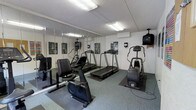 This single-floor fitness room is designed to provide a practical and well-equipped workout space. The room features a selection of cardio equipment, including treadmills, a stationary bike, and an elliptical cross-trainer, all arranged to allow comfortable use. A full wall of mirrors lines one side of the room, creating a sense of expanded space while allowing users to monitor their form during exercise. The flooring consists of durable, rubber-backed exercise matting beneath the equipment, set against a grey utility-style floor that is built to withstand heavy use. Overhead, flush-mounted fluorescent lighting ensures the room is well-lit throughout. A wall-mounted television is positioned for easy viewing from the cardio machines, adding an entertainment element to workouts. A wall-mounted air conditioning unit provides climate control, keeping the environment comfortable year-round. Motivational fitness posters are displayed along the walls, contributing to the gym's energetic atmosphere. The textured ceiling and clean white walls give the room a straightforward, no-frills aesthetic focused entirely on functionality. Overall, this fitness room offers a complete and well-maintained environment suited for regular cardiovascular training.