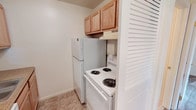 This single-floor unit features a straightforward, functional layout with a neutral color palette of white walls and beige carpet throughout the living and sleeping areas. The spacious main living area is anchored by a ceiling fan with integrated lighting and includes a wall-mounted sconce near the entry door, providing ambient illumination. A baseboard heating system runs along the perimeter walls, offering consistent climate control. A closet is accessible directly from the living space, adding practical storage.

The kitchen is efficiently arranged, featuring white coil-burner electric range, a full-size refrigerator, and a range hood overhead. Light wood upper cabinetry provides storage, while the flooring transitions to tile for easy maintenance.

The bedroom offers comfortable proportions with wall-to-wall carpet, a ceiling fan with light fixture, a window allowing natural light, and a baseboard heater along one wall. An additional room with carpet and baseboard heating provides flexible space that could serve various purposes.

The bathroom is clean and simply appointed, featuring a full-sized soaking tub with an overhead shower, white square tile surround, and chrome fixtures. Overall, the unit presents a practical, no-frills living environment with consistent finishes throughout.