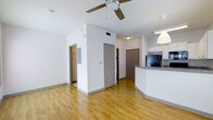 This single-floor unit showcases an open-concept layout that seamlessly connects the living area to the kitchen. The main living space is finished with warm hardwood-style flooring that extends throughout, complementing the clean white walls and adding a sense of continuity across the unit. A ceiling fan is centrally mounted overhead, providing both airflow and a functional lighting element.

The kitchen is positioned along one side of the open area and features a curved granite countertop that doubles as a breakfast bar, white cabinetry, and black appliances including a refrigerator. A pendant light hangs above the counter, adding a focused lighting accent to the space. The kitchen layout allows for easy interaction with the living area, making the space well-suited for everyday use.

Multiple interior doors lead to additional areas within the unit, including what appears to be a utility or storage closet housing an electrical panel. A thermostat is mounted on a central wall column, offering convenient climate control access. The overall design is straightforward and functional, with neutral tones and practical finishes throughout.