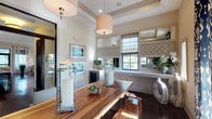 This single-story space showcases a thoughtfully designed dining area that masterfully balances natural warmth with contemporary sophistication. The centerpiece is a stunning live-edge wood dining table, whose organic form and rich grain patterns command immediate attention. Its natural character is complemented by an eclectic mix of seating, including sculptural open-lattice chairs and upholstered dining chairs.

The elevated ceilings feature tray detailing and are adorned with multiple drum pendant lights in neutral linen tones, casting a warm, inviting glow over the space. Dark hardwood flooring grounds the room and provides striking contrast against the crisp white walls and wainscoting.

A built-in sideboard with a beverage refrigerator lines one wall, topped with a mirror framed by a decorative geometric lattice panel, adding depth and visual interest. Multiple windows allow natural light to flow in, offering outdoor views, and are dressed with Roman shades and bold navy ikat-patterned drapes. Glass lantern candle holders and curated coastal-inspired artwork complete the refined, resort-like ambiance throughout.