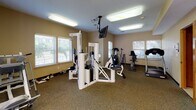 This fitness center is organized across a single floor and divided into distinct workout zones, offering a comprehensive range of exercise options. The main cardio area is spacious and well-lit, featuring multiple windows with slatted blinds that allow natural light to fill the room. Equipment here includes a treadmill, stationary bike, elliptical, and a recumbent bike, alongside a ceiling-mounted television for entertainment during workouts. Flooring throughout this zone consists of durable, low-pile carpet.

A large multi-station cable machine anchors the center of the main room, providing options for a variety of resistance exercises. Adjacent to the cardio area, a dedicated free weights section features rubber mat flooring for safety and noise reduction. This zone is equipped with a full dumbbell rack, a weight bench with barbell attachments, and additional strength training equipment. A wall-mounted mirror in this area enhances the sense of space and assists with form.

Near the entrance, a water cooler and accessible storage are conveniently positioned. The neutral beige walls, recessed and flush-mounted ceiling lighting, and multiple entry points contribute to a clean, functional, and welcoming training environment throughout the facility.