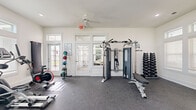 This fitness center offers a comprehensive workout facility within a clean, modern space. The room is finished with durable speckled rubber flooring throughout, providing both safety and functionality for a wide range of exercises. Bright white walls and multiple windows with transom details allow natural light to fill the space, creating an open and energizing atmosphere. A ceiling fan overhead aids in air circulation during workouts.

The cardio zone features two Spirit treadmills, an elliptical trainer, and a stationary spin bike, all positioned along a windowed wall for an airy feel. A wall-mounted flat-screen television provides entertainment during workouts. On the strength training side, the facility includes a multi-function cable machine with an adjustable bench, a full rack of dumbbells, medicine balls on a vertical storage rack, and a barbell with weight plates. Two interior doors lead to additional rooms, likely restrooms or utility spaces. French-style glass doors with transom windows serve as the main entry, connecting to an adjacent common area. The overall design is clean, bright, and functional, making it a well-rounded fitness facility suited for both cardio and strength training needs.