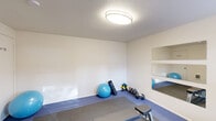 Located at 5817 SW Beaverton Hillsdale Hwy in Portland, Oregon, this single-floor fitness facility offers a well-organized and functional space designed for a variety of workout routines. The facility is divided into two exercise rooms, each serving a distinct purpose, complemented by a dedicated accessible bathroom.

The primary cardio room features durable rubber flooring and is equipped with a treadmill, a recumbent exercise bike, and an upright stationary bike. A large sliding glass door with vertical blinds floods the room with natural light and provides views of the surrounding greenery, creating an open and inviting atmosphere. A round flush-mount ceiling light provides additional illumination.

The second exercise room is configured for strength and flexibility training, with interlocking foam and rubber floor tiles providing a safe workout surface. The room is outfitted with a weight bench, a dumbbell rack, stability balls, a foam roller, and wall-mounted mirrors that enhance the sense of space. A window with horizontal blinds allows natural light to enter, keeping the room bright and comfortable.

The accessible bathroom features wood-look luxury vinyl flooring, grab bars, a wall-mounted paper towel dispenser, and tasteful wall art, ensuring convenience and accessibility for all users. Clean, neutral wall tones throughout the facility contribute to a calm, focused environment suitable for daily exercise.