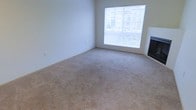 This single-floor space offers a clean, functional layout designed to maximize comfort and livability. The main living area is a standout feature, characterized by generous proportions and soft, wall-to-wall carpeting in a neutral beige tone that adds warmth underfoot. A large picture window dominates one wall, flooding the room with an abundance of natural light and offering views of the surrounding urban environment, including neighboring buildings and greenery. The bright, white walls enhance the sense of openness and provide a versatile backdrop for a variety of interior styles. A sleek fireplace is positioned along the adjacent wall, serving as a functional focal point that contributes both visual interest and a cozy atmosphere. Electrical outlets are conveniently placed throughout the room, ensuring practical usability. The overall ambiance of the space is light, airy, and move-in ready, with a neutral palette that accommodates a wide range of furnishing and décor choices. The single-floor layout promotes ease of movement and accessibility throughout the unit, making it a straightforward and efficient living environment.