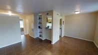 This single-story space features an open, flowing layout anchored by warm terra cotta tile flooring throughout. A central structural column divides the main living area, incorporating a set of built-in display shelves on one side—ideal for books, décor, or personal items. A pass-through opening connects the living area to the kitchen, which is equipped with light wood cabinetry, white appliances, and a countertop that doubles as a casual bar or serving surface. The kitchen is also accessible via a dedicated entryway corridor, which includes a mounted fire extinguisher and leads further into the unit. The living area is generously proportioned, with neutral beige walls and a textured ceiling that complement the earthy tones of the flooring. A wall-mounted HVAC vent and utility panel are integrated discreetly into the living space. Natural light enters through a window visible at the far end of the hallway, adding brightness to the otherwise warmly lit interior. The overall ambiance is functional and practical, with a straightforward layout that maximizes usable floor space across the single level.