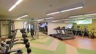 This fitness center offers a comprehensive workout environment designed to accommodate a wide range of exercise needs. The space is bathed in bright, even illumination provided by rows of linear pendant and recessed ceiling lights, creating a clean and energizing atmosphere throughout.

The cardio zone features a lineup of modern treadmills and additional cardio machines, positioned to face a large wall-mounted flat-screen television — keeping exercisers entertained during their sessions. Adjacent to the cardio area, a dedicated strength training zone houses a multi-station cable and weight machine alongside a robust free weight section, complete with a full dumbbell rack offering a wide range of weights.

The flooring is a standout design element, featuring a bold geometric pattern in alternating green and neutral tones that visually separates workout zones while adding vibrancy to the space. The walls are finished in a clean, neutral white, with mirrored panels strategically placed to expand the sense of space and allow users to monitor their form during exercise.

The layout is thoughtfully organized to maximize usability, with clear pathways between equipment zones ensuring safe and efficient movement throughout the facility. Overall, this fitness center combines practical functionality with a polished, modern aesthetic.