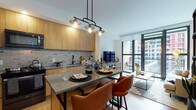 This single-floor residence is defined by its contemporary design and abundant natural light. The open-plan living and kitchen area serves as the heart of the space, where large floor-to-ceiling windows frame sweeping urban views, flooding the interior with daylight. Light oak hardwood flooring runs throughout, creating a warm, cohesive foundation across all rooms.

The kitchen is thoughtfully designed with warm wood lower cabinetry, crisp white upper cabinets, and a marble-style tile backsplash. Stainless steel appliances — including a range, microwave, refrigerator, and dishwasher — complement the clean white countertops, balancing functionality with modern aesthetics.

The living area offers a generous layout accommodating comfortable seating and an entertainment setup, all oriented toward the expansive city-facing windows. A dedicated dining nook sits nearby, featuring a round wood table with mid-century modern chairs beneath a crisp white wall adorned with framed artwork.

The bedroom continues the bright, airy theme with large windows offering urban views, light hardwood floors, and clean white walls. The overall ambiance throughout the space is modern, light-filled, and urban, with a consistent mid-century modern aesthetic.