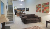 This two-story space makes an immediate impression with its dramatically high ceilings in the main living area, creating a sense of openness and volume that defines the overall ambiance. The open-concept layout seamlessly connects the living room to the kitchen and dining area, promoting a fluid, social atmosphere. The living area is anchored by plush, dark brown upholstered seating arranged around a patterned area rug, complemented by warm lamp lighting and colorful wall art that adds a welcoming, casual character. A side table provides functional surface space alongside the seating arrangement. The kitchen area features wood-toned cabinetry and a breakfast bar with counter seating, making it well-suited for both meal preparation and casual dining. A dining table with chairs sits just adjacent, creating a defined eating space within the open floor plan. Chalkboard signage adds a personalized, informal touch to the kitchen wall. Light-toned carpeting runs throughout the main level, contributing to the warm and relaxed feel of the space. With two floors, the layout offers additional functional space beyond the main living level.