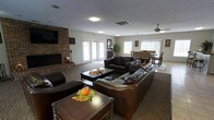 This single-floor space is designed to offer a warm and inviting atmosphere for gathering and relaxation. The main lounge area is anchored by a classic full-height brick fireplace, above which a flat-screen television is mounted, creating a natural focal point for the room. Rich, dark leather sofas and armchairs are arranged around a wooden coffee table, providing ample and comfortable seating throughout the space. The patterned carpet adds texture and warmth underfoot, complementing the earthy tones of the décor. Recessed and flush-mount ceiling lighting ensures the space is well-lit, while multiple windows along the walls allow natural light to filter in, brightening the interior. A ceiling fan aids in air circulation, adding to the overall comfort of the environment. Adjacent to the lounge, a dedicated dining area features a dark wood table and matching chairs, ideal for shared meals or group activities. Lush potted greenery adds a touch of nature to the space. French-style doors offer access to the exterior, further enhancing the connection between indoor and outdoor areas. Overall, the space exudes a comfortable, community-oriented ambiance suited for socializing and everyday use.