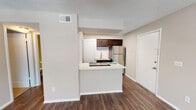 This single-story space is thoughtfully laid out, combining functional design with modern finishes throughout. The main living area is bright and open, featuring rich dark hardwood-style flooring, neutral-toned walls, a textured ceiling, and a ceiling fan with integrated lighting. A wide window fitted with horizontal blinds allows generous natural light to fill the room, while a glass-panel door provides additional access and further brightens the space.

The kitchen is efficiently designed in a peninsula configuration, offering a white countertop surface that extends into a breakfast bar for casual dining. Dark espresso-toned cabinetry provides ample storage, complemented by a white subway tile backsplash, a stainless steel refrigerator, a built-in cooktop, and a stainless steel sink. A skylight above the kitchen adds additional brightness. An in-unit stacked washer and dryer are conveniently located nearby.

The bedroom is carpeted for comfort and includes its own ceiling fan with light and a window that brings in natural light. The full bathroom is well-appointed with a dark-framed vanity mirror, espresso-toned cabinetry, a white countertop, a single sink, and a tub-shower combination. Dark hardwood-style flooring carries through the bathroom, maintaining design consistency throughout the unit.