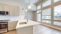 This single-floor residence offers a clean, modern aesthetic defined by a neutral color palette, light hardwood flooring, and thoughtfully designed living spaces. The open-concept kitchen serves as the centerpiece of the home, showcasing a large peninsula with elegant marble-veined quartz countertops and an undermount stainless steel sink. Geometric pendant lights hang above the peninsula, adding a contemporary touch. Taupe shaker-style cabinetry lines the kitchen walls, complemented by a crisp white tile backsplash and a full suite of stainless steel appliances, including a gas range, over-the-range microwave, refrigerator, and dishwasher. The kitchen opens seamlessly to a living area bathed in natural light from floor-to-ceiling windows, which offer views to an adjacent outdoor space. The elevated ceilings throughout enhance the sense of openness and airiness. The bathroom features a soaking tub and shower combination framed by large-format, cream-toned stone tile, with chrome fixtures that add a refined finish. Additional highlights include a generously sized walk-in closet outfitted with multiple shelving rods and hardwood flooring, providing ample storage. Together, these elements create a bright, well-organized, and stylish living environment.