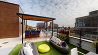 This rooftop terrace offers a well-appointed outdoor living experience elevated above a bustling urban skyline. The space is designed for both relaxation and entertaining, featuring distinct zones that cater to a variety of uses. A lush artificial turf area anchors the lounge section, where contemporary pod-style seating and modular chairs are arranged around a bright chartreuse ottoman, creating a casual and inviting gathering area. Bold red ornamental plants in large planters line the perimeter, adding striking color contrast against the city backdrop.

A covered pergola structure with warm wood-paneled ceiling and sleek black steel framing defines a dedicated bar and dining area. This sheltered zone features a built-in bar with blue upholstered high stools and an adjacent dining table with white chairs, making it ideal for hosting. Glass railings along the terrace edges preserve unobstructed panoramic views of the surrounding cityscape, including high-rise buildings and expansive open sky. The overall ambiance is modern and vibrant, blending sophisticated design elements with a lively urban atmosphere.