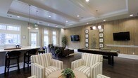 This single-floor communal space is thoughtfully designed to serve as a versatile social hub. The interior is anchored by a tray ceiling with recessed lighting and elegant pendant fixtures, lending the room a polished, upscale feel. Dark hardwood flooring runs throughout, providing a warm contrast to the light-toned walls and furnishings.

The seating area in the foreground features striped upholstered armchairs arranged around a central coffee table, creating an inviting conversation space. Behind it, a second lounge zone offers dark leather sofas centered around a wall-mounted flat-screen television set against a wood-paneled accent wall. The panel wall adds texture and warmth to the overall design.

To the right, a full-size pool table is positioned beneath a row of pendant lights, offering a dedicated recreation area. A gallery of framed artwork lines the walls nearby, adding a curated, refined touch.

Large windows dressed with sheer curtains allow ample natural light to fill the space, while potted plants are placed throughout to introduce natural elements. The overall ambiance balances comfort and sophistication, making it well-suited for both quiet relaxation and casual social gatherings.