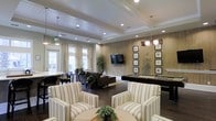 This single-floor communal space is thoughtfully designed to serve as a versatile social hub. The interior is anchored by a tray ceiling with recessed lighting and elegant pendant fixtures, lending the room a polished, upscale feel. Dark hardwood flooring runs throughout, providing a warm contrast to the light-toned walls and furnishings.

The seating area in the foreground features striped upholstered armchairs arranged around a central coffee table, creating an inviting conversation space. Behind it, a second lounge zone offers dark leather sofas centered around a wall-mounted flat-screen television set against a wood-paneled accent wall. The panel wall adds texture and warmth to the overall design.

To the right, a full-size pool table is positioned beneath a row of pendant lights, offering a dedicated recreation area. A gallery of framed artwork lines the walls nearby, adding a curated, refined touch.

Large windows dressed with sheer curtains allow ample natural light to fill the space, while potted plants are placed throughout to introduce natural elements. The overall ambiance balances comfort and sophistication, making it well-suited for both quiet relaxation and casual social gatherings.