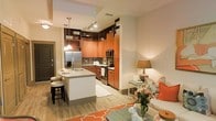 This single-floor residence showcases a thoughtfully designed open-concept layout that seamlessly connects the kitchen and living areas. The kitchen is a standout feature, boasting rich warm-toned wood cabinetry that extends to the upper walls, maximizing storage with tall upper cabinets and a built-in wine rack. A central island with a granite countertop and undermount sink doubles as a breakfast bar with bar stool seating, offering both functionality and a social gathering point. Stainless steel appliances, including a refrigerator, range, and microwave, complement the mosaic tile backsplash, adding a contemporary touch. Track lighting and pendant lights illuminate the kitchen efficiently. The living area flows naturally from the kitchen, featuring light hardwood-style flooring that runs throughout the space, creating a cohesive aesthetic. Warm, neutral wall tones are accented by vibrant orange decor elements, lending energy and character to the interior. The overall ambiance is modern yet inviting, with layered lighting and an open layout that enhances the sense of spaciousness throughout the unit.