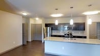This single-floor space showcases a well-designed open-concept layout centered around a contemporary kitchen. The kitchen is outfitted with dark espresso-toned cabinetry that contrasts cleanly against light granite countertops, which extend across a spacious central island complete with an undermount sink and modern faucet. Two sleek pendant lights hang above the island, complementing the flush-mount ceiling fixtures that provide consistent, even illumination throughout the space.

Stainless steel appliances, including a refrigerator, electric range, and built-in microwave, add a polished, cohesive aesthetic. A subtle tile backsplash runs along the wall behind the range, adding texture and visual interest. The dark hardwood-style flooring grounds the space with warmth and continuity, flowing seamlessly from the kitchen into the adjoining areas.

The open sightlines from the kitchen reveal a hallway leading to multiple doors, suggesting well-separated rooms beyond. The neutral wall tones paired with the dark cabinetry and flooring create a balanced, contemporary ambiance. Overall, the space presents a functional yet refined design with thoughtful finishes throughout.