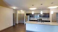 This single-floor space showcases a well-designed open-concept layout centered around a contemporary kitchen. The kitchen is outfitted with dark espresso-toned cabinetry that contrasts cleanly against light granite countertops, which extend across a spacious central island complete with an undermount sink and modern faucet. Two sleek pendant lights hang above the island, complementing the flush-mount ceiling fixtures that provide consistent, even illumination throughout the space.

Stainless steel appliances, including a refrigerator, electric range, and built-in microwave, add a polished, cohesive aesthetic. A subtle tile backsplash runs along the wall behind the range, adding texture and visual interest. The dark hardwood-style flooring grounds the space with warmth and continuity, flowing seamlessly from the kitchen into the adjoining areas.

The open sightlines from the kitchen reveal a hallway leading to multiple doors, suggesting well-separated rooms beyond. The neutral wall tones paired with the dark cabinetry and flooring create a balanced, contemporary ambiance. Overall, the space presents a functional yet refined design with thoughtful finishes throughout.